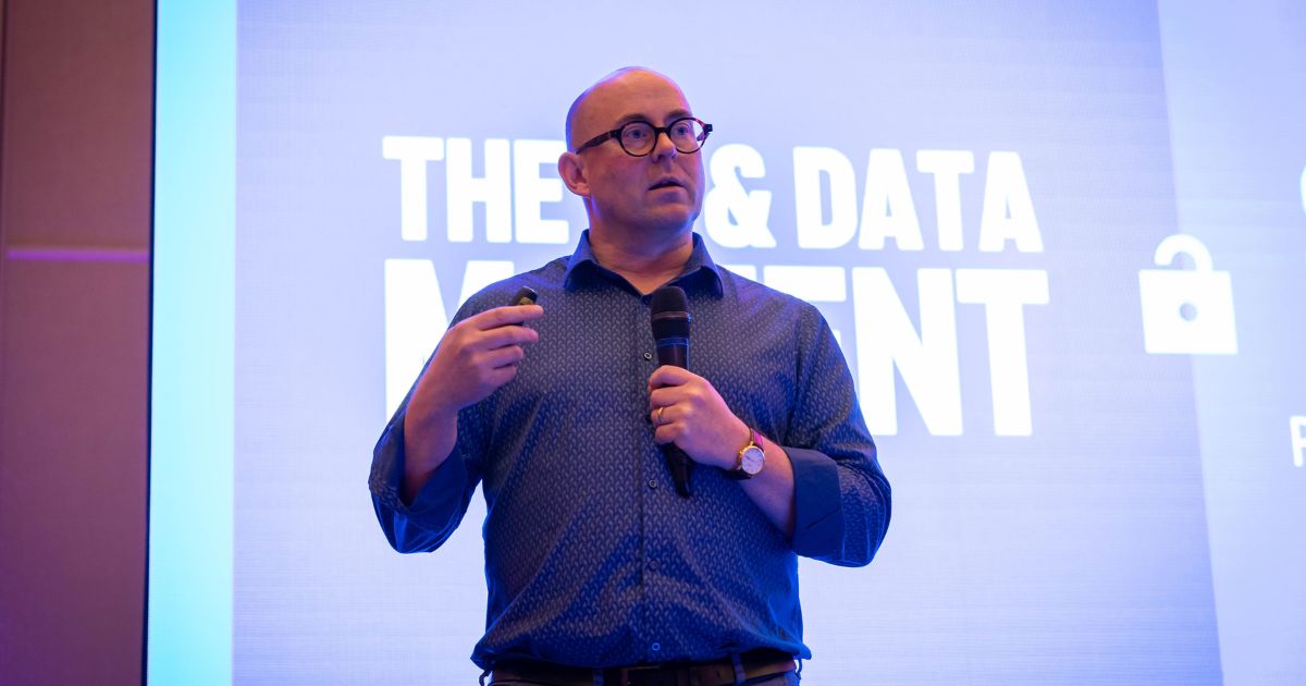 A imagem mostra uma pessoa em pé, aparentemente realizando uma apresentação ou palestra. A cena inclui os seguintes elementos: Contexto geral: Ambiente interno, provavelmente uma sala de conferências ou auditório, com iluminação artificial. O fundo é uma tela grande com projeção de texto. Texto visível no fundo: “THE & DATA MENT” (provavelmente parte de uma frase maior como “THE DATA MANAGEMENT” ou similar). Há também um ícone de cadeado à direita, sugerindo um tema relacionado à segurança ou proteção de dados. Pessoa: Está vestindo uma camisa social azul com padrão discreto e mangas dobradas. Usa um relógio no pulso esquerdo e segura dois objetos: um microfone na mão direita e um controle remoto ou apontador na mão esquerda, indicando que está conduzindo a apresentação. Cores e iluminação: Predominância de tons azuis e roxos, com luz forte vinda da tela projetada, criando um contraste com a figura em primeiro plano. (NetApp)