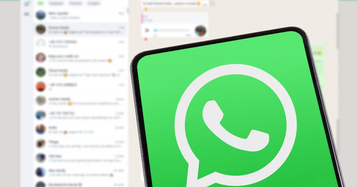 A imagem mostra um smartphone com a tela exibindo o ícone do WhatsApp em destaque, sobre um fundo que parece ser a interface do aplicativo aberta em um computador ou versão web. Detalhes principais: Primeiro plano: Ícone do WhatsApp em verde com o símbolo branco característico (balão de conversa com telefone). O enquadramento foca no dispositivo, ocupando a maior parte da imagem. Plano de fundo: Interface do WhatsApp Web ou Desktop, com lista de conversas visível à esquerda e uma conversa aberta à direita. Elementos como nomes, mensagens e emojis aparecem desfocados, mantendo a privacidade. Cores e iluminação: Predominância de tons verdes e brancos, reforçando a identidade visual do WhatsApp. Iluminação clara e uniforme, criando uma composição limpa e moderna. Essa imagem transmite comunicação digital, conectividade e integração entre dispositivos móveis e desktop.
