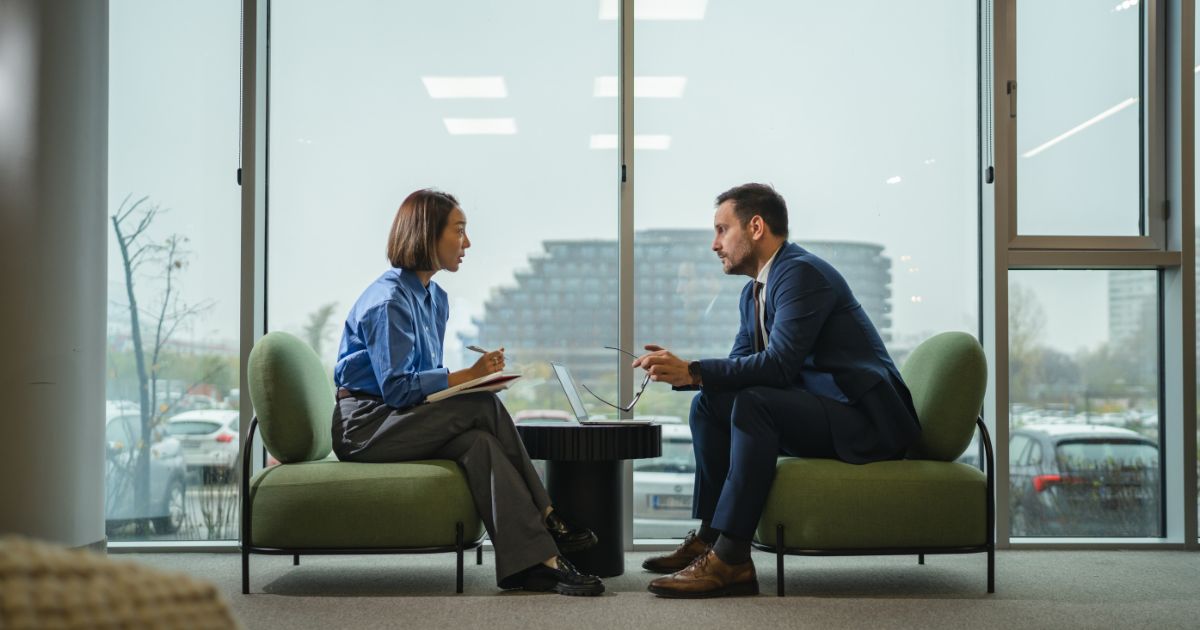 A imagem mostra duas pessoas em uma reunião profissional, sentadas frente a frente em poltronas verdes, diante de uma grande janela de vidro que oferece vista para um ambiente urbano com edifícios ao fundo. Descrição da cena Ambiente: Espaço corporativo moderno, com ampla iluminação natural graças às janelas do chão ao teto. O fundo mostra carros estacionados e prédios, sugerindo que a sala está localizada em um edifício comercial. Interação: Uma pessoa está com um bloco de anotações, em postura ativa de escuta, sugerindo entrevista, reunião de alinhamento ou acompanhamento profissional. A outra pessoa, vestindo um terno azul, segura óculos na mão, em posição inclinada para frente, indicando atenção e diálogo direto. Entre eles, há um laptop aberto sobre uma pequena mesa, reforçando o contexto corporativo e colaborativo. Clima geral: A cena transmite profissionalismo, troca de informações e um ambiente de negócios acolhedor. A composição sugere um momento de tomada de decisão, mentoria, entrevista ou negociação. (trabalho)