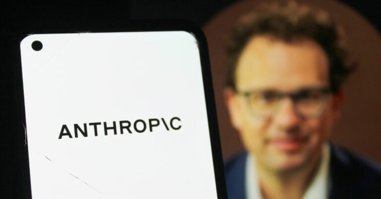 Telefone celular exibindo, em destaque, o nome “ANTHROPIC” em letras pretas sobre fundo branco. Ao fundo, aparece uma pessoa usando roupas formais, em segundo plano, com iluminação suave e foco concentrado no dispositivo móvel. (humanidade)