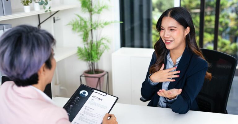 A imagem mostra uma cena de entrevista de emprego em um ambiente corporativo. No primeiro plano, há uma pessoa segurando uma prancheta com um currículo visível, contendo a palavra “RESUME” no topo e informações organizadas em seções. Do outro lado da mesa, outra pessoa está gesticulando com uma das mãos sobre o peito, sugerindo que está respondendo a perguntas ou explicando algo. Ambas estão sentadas em cadeiras, e a mesa é branca, com um ambiente moderno ao fundo, incluindo uma janela ampla com vista para áreas verdes e prateleiras com objetos decorativos. A composição transmite um contexto profissional, relacionado a recrutamento, seleção ou entrevista de trabalho.