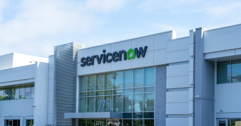 Fachada de um prédio corporativo moderno com arquitetura contemporânea, composta por grandes painéis de vidro e revestimento em tons claros. No topo do edifício, aparece o logotipo “ServiceNow”, com a palavra “Service” em preto e “Now” em verde. O céu azul e a iluminação natural destacam o ambiente empresarial e tecnológico.
