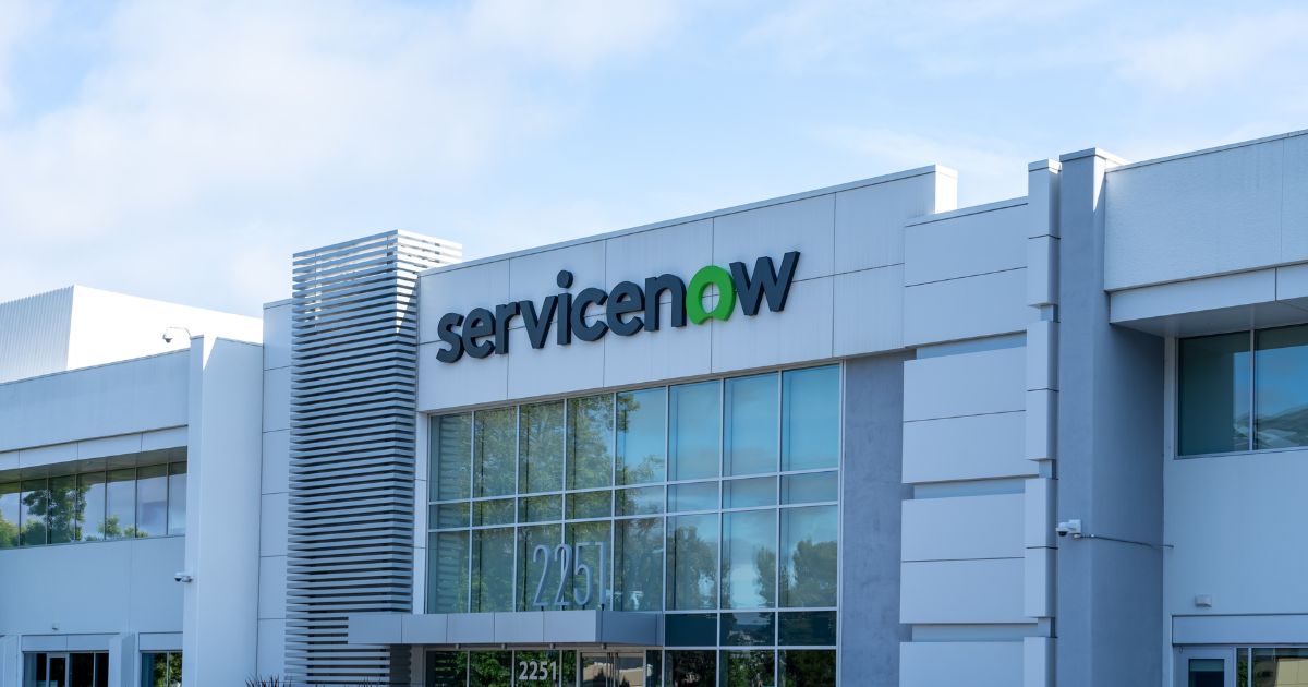 Fachada de um prédio corporativo moderno com arquitetura contemporânea, composta por grandes painéis de vidro e revestimento em tons claros. No topo do edifício, aparece o logotipo “ServiceNow”, com a palavra “Service” em preto e “Now” em verde. O céu azul e a iluminação natural destacam o ambiente empresarial e tecnológico.