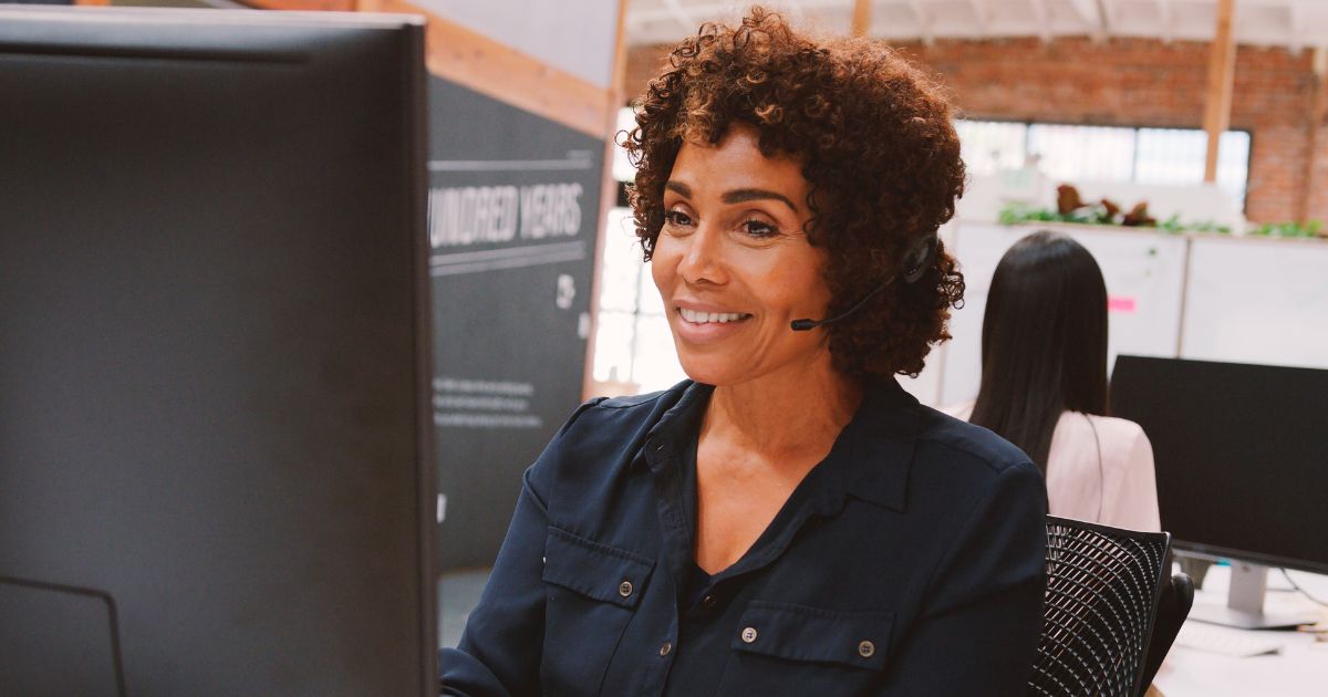 A imagem mostra um ambiente de escritório moderno com duas pessoas trabalhando em frente a computadores. Detalhes principais: Primeiro plano: Uma pessoa está usando um headset e olhando para um monitor grande, sugerindo atividades como atendimento ao cliente, suporte técnico ou videoconferência. A pessoa veste uma camisa escura com bolsos frontais. Segundo plano: Outra pessoa está sentada em frente a um computador, com cabelos longos e lisos, vestindo uma peça clara. Ambiente: O espaço tem um design industrial, com paredes de tijolos aparentes e vigas de madeira no teto. Há divisórias baixas e algumas plantas decorativas, criando um clima de trabalho colaborativo e acolhedor. Iluminação: Natural e suave, provavelmente proveniente de janelas amplas, reforçando a sensação de um espaço aberto e bem iluminado. (clientes)