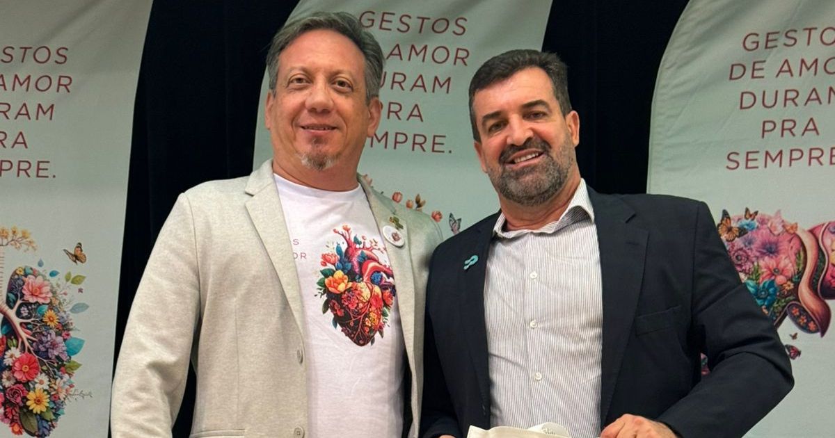 A imagem mostra duas pessoas lado a lado em um evento institucional com uma estética acolhedora e humanizada. Elementos principais da cena Vestuário: Uma pessoa veste um blazer claro com uma camiseta ilustrada com um coração composto por elementos florais e naturais, criando uma conexão visual com o tema da campanha. A outra veste um blazer escuro com camisa social clara, trazendo um tom mais formal ao conjunto. Ambiente: Ao fundo, há grandes banners verticais com a frase: “GESTOS DE AMOR DURAM PRA SEMPRE”. Os banners também exibem ilustrações vibrantes com flores, borboletas e elementos orgânicos, reforçando a mensagem de cuidado, vida e afeto. Clima da imagem: A cena transmite união, colaboração e participação em uma iniciativa com foco em solidariedade, conscientização ou responsabilidade social. O visual geral é caloroso e marcante, com cores vívidas e elementos gráficos simbólicos. (transplante, órgãos, Paipe)