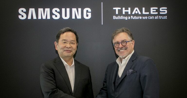 Duas pessoas usando trajes formais apertam as mãos diante de um painel preto com os logotipos “Samsung” e “Thales” acompanhados do slogan “Building a future we can all trust”. A cena representa uma parceria corporativa ou assinatura de acordo entre as duas empresas.