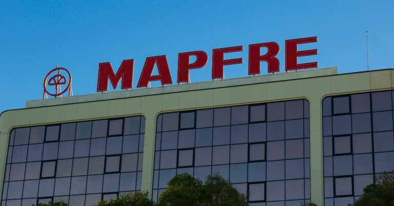 A imagem mostra a fachada de um edifício corporativo com o logotipo e nome MAPFRE em destaque na parte superior. Detalhes principais: Texto visível: “MAPFRE” em letras grandes e vermelhas, acompanhado do símbolo da empresa à esquerda. Arquitetura: Estrutura moderna com linhas retas e revestimento claro. Fileiras de janelas espelhadas, refletindo o céu e parte da vegetação ao redor. Ambiente: Céu limpo e azul, indicando um dia ensolarado. Vegetação na base da imagem, sugerindo árvores próximas ao prédio. Composição visual: O contraste entre o vermelho do logotipo e o azul do céu cria uma imagem marcante e profissional. Essa imagem remete a instituições financeiras, seguros e soluções corporativas, transmitindo solidez e credibilidade.