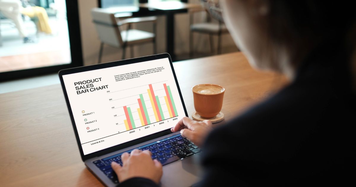 Pessoa sentada diante de um notebook em uma mesa de madeira, analisando um gráfico de barras intitulado “Product Sales Bar Chart”, que mostra comparações de vendas entre os anos de 2020 e 2023. Ao lado do computador, há uma xícara de café em um pires. O ambiente parece ser um café ou espaço de trabalho com móveis claros e janelas amplas ao fundo. (vendas)