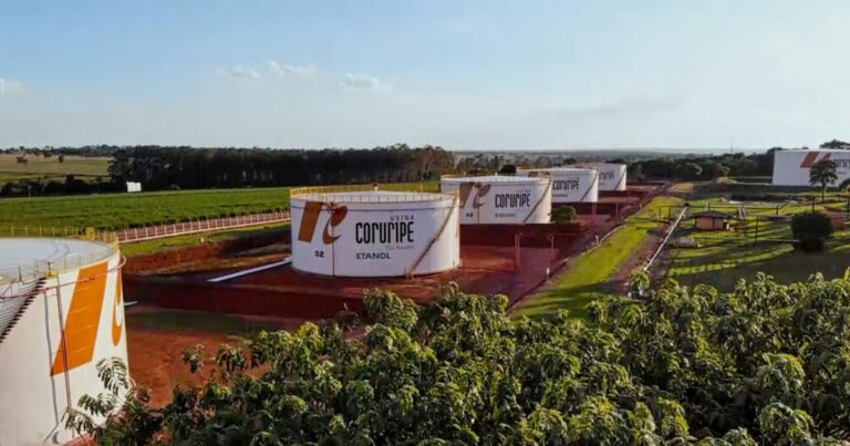 A imagem mostra uma área industrial ao ar livre, composta por diversos tanques cilíndricos de grande porte utilizados para armazenamento. Cada tanque é pintado de branco com detalhes em laranja e exibe a marca “Usina Coruripe” e a palavra “Etanol”. Os tanques estão distribuídos ao longo de um terreno amplo, com solo avermelhado e caminhos internos organizados. Ao redor da instalação, há extensas áreas verdes, incluindo plantações e áreas de vegetação baixa. No horizonte, é possível ver faixas de árvores e o céu claro de fim de tarde, iluminando toda a paisagem com luz natural suave.