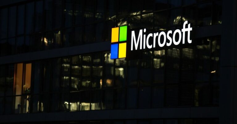 A imagem mostra a fachada de um edifício corporativo com o logotipo da Microsoft iluminado em destaque. Detalhes principais: Texto visível: “Microsoft” escrito em branco, acompanhado do ícone característico da empresa — um quadrado dividido em quatro partes coloridas (vermelho, verde, azul e amarelo). Arquitetura: Estrutura moderna com grandes painéis de vidro refletindo luzes internas e externas. Iluminação noturna, com luzes acesas dentro do prédio, criando um contraste elegante com o fundo escuro. Ambiente: A cena sugere um horário após o expediente ou início da noite, transmitindo uma atmosfera corporativa e tecnológica. Essa imagem remete à inovação, tecnologia e presença global da Microsoft, reforçando sua identidade como líder em soluções digitais.