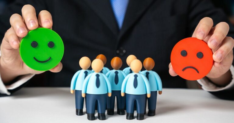 Pessoa vestindo terno segura um círculo verde com um rosto sorridente na mão esquerda e um círculo vermelho com um rosto triste na mão direita. À frente, há pequenos bonecos que representam um grupo de pessoas, todos com roupas formais em azul. A cena simboliza avaliação ou classificação positiva e negativa. (RH)