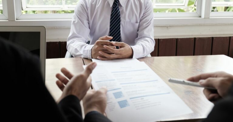 A imagem mostra uma cena típica de entrevista ou reunião profissional em um ambiente corporativo. Detalhes principais: Primeiro plano: Duas mãos segurando um currículo ou documento impresso, com texto visível em formato de lista, indicando que é provavelmente um CV ou proposta. Outra mão segura uma caneta, sugerindo anotações ou avaliação. Centro da imagem: Pessoa vestindo camisa social branca e gravata listrada azul, com as mãos entrelaçadas sobre a mesa, postura formal e atenta. Ambiente: Mesa de madeira clara, com um laptop parcialmente visível à esquerda. Fundo com janelas amplas, permitindo entrada de luz natural, e plantas do lado externo, criando um clima profissional e acolhedor. Essa imagem transmite claramente processos seletivos, entrevistas de emprego ou reuniões de negócios.