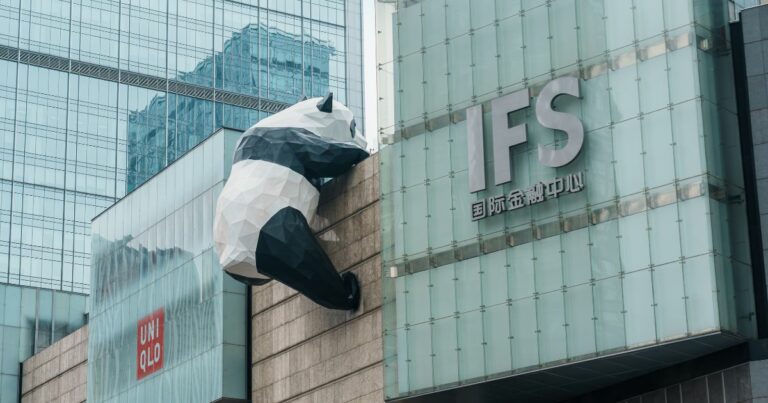 Fachada de um edifício moderno com paredes de vidro, onde se destaca uma grande escultura de panda em estilo geométrico, posicionada como se estivesse escalando a parede. À direita, aparece o logotipo “IFS” acompanhado de caracteres asiáticos. Ao fundo, outros prédios altos refletem-se nas superfícies envidraçadas, compondo um cenário urbano contemporâneo.