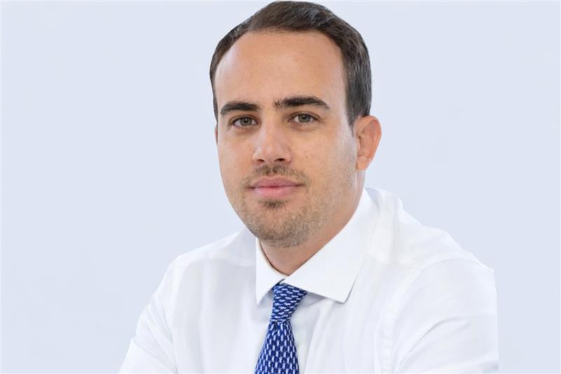 Pessoa vestindo uma camisa social branca e uma gravata azul com padrão discreto, sentada diante de um fundo claro e uniforme. A pose é formal, sugerindo um retrato profissional ou corporativo. (telemedicina)