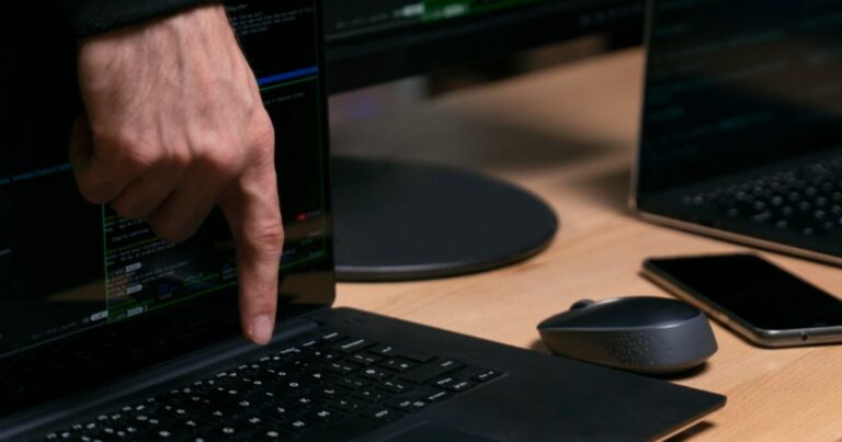 Uma mão aponta para o teclado de um laptop enquanto várias linhas de código são exibidas na tela. O computador está sobre uma mesa de madeira, ao lado de um mouse cinza, um smartphone e outro laptop parcialmente visível. Ao fundo, há monitores adicionais mostrando interfaces de programação, criando um ambiente típico de trabalho com desenvolvimento de software ou análise técnica. (Caso Epstein)