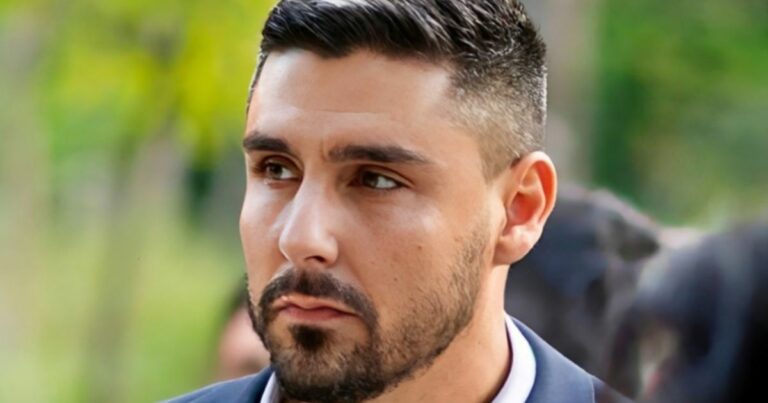 Uma pessoa com cabelo curto e escuro, bem arrumado, veste um traje formal em um ambiente ao ar livre. O fundo apresenta vegetação desfocada em tons de verde e amarelo, sugerindo um parque ou área natural iluminada. Outras pessoas aparecem suavemente ao fundo, também desfocadas, indicando que a cena pode ser parte de um evento social ou cerimônia ao ar livre. (ANTT)