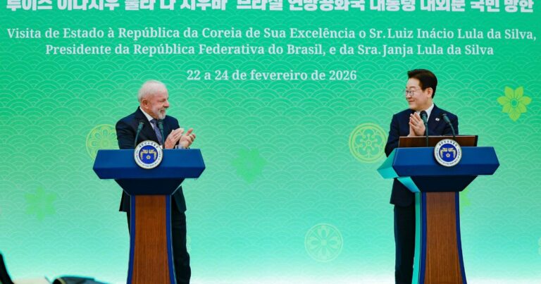 Dois representantes oficiais estão em pé atrás de púlpitos em um palco com fundo verde decorado com elementos gráficos e texto bilíngue em coreano e português. Eles aplaudem um ao outro em um ambiente formal de cerimônia diplomática. No painel ao fundo, há informações sobre uma visita de Estado à Coreia do Sul, incluindo datas e nomes das autoridades envolvidas. (Coreia do Sul)