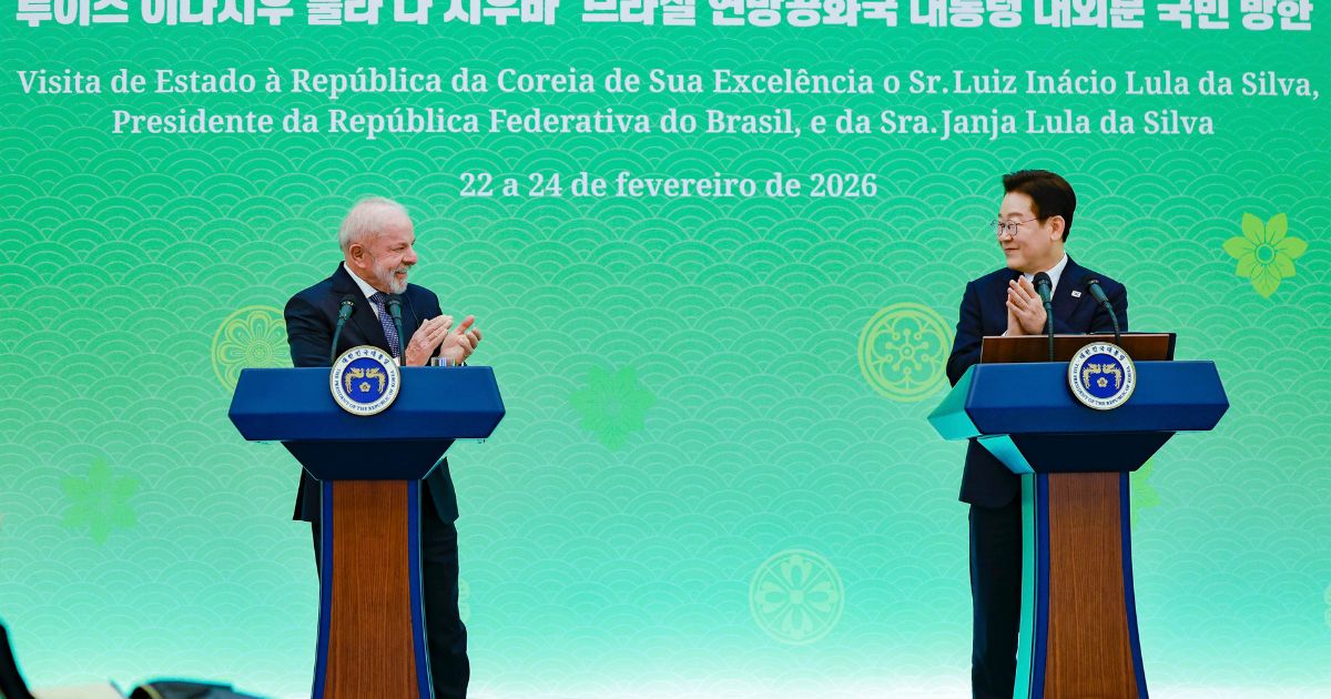 Dois representantes oficiais estão em pé atrás de púlpitos em um palco com fundo verde decorado com elementos gráficos e texto bilíngue em coreano e português. Eles aplaudem um ao outro em um ambiente formal de cerimônia diplomática. No painel ao fundo, há informações sobre uma visita de Estado à Coreia do Sul, incluindo datas e nomes das autoridades envolvidas. (Coreia do Sul)