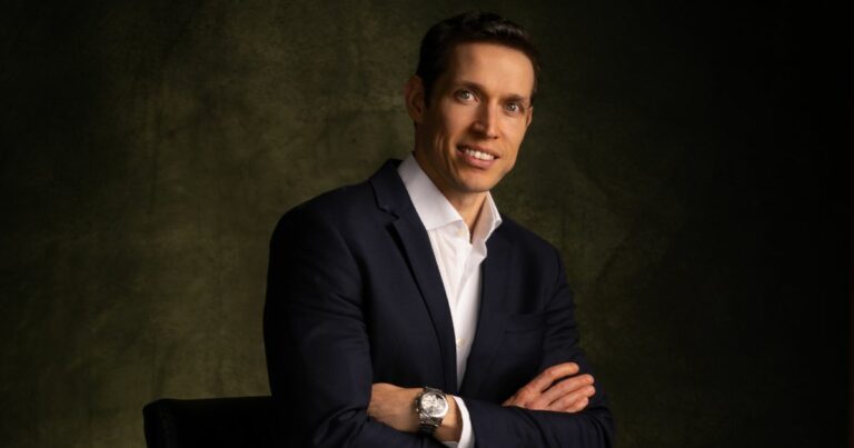 Pessoa vestindo um terno azul-escuro com camisa social branca, posando com os braços cruzados. A iluminação é dramática, destacando a silhueta e o relógio metálico no pulso esquerdo, diante de um fundo escuro com textura suave. (Bureau Veritas)