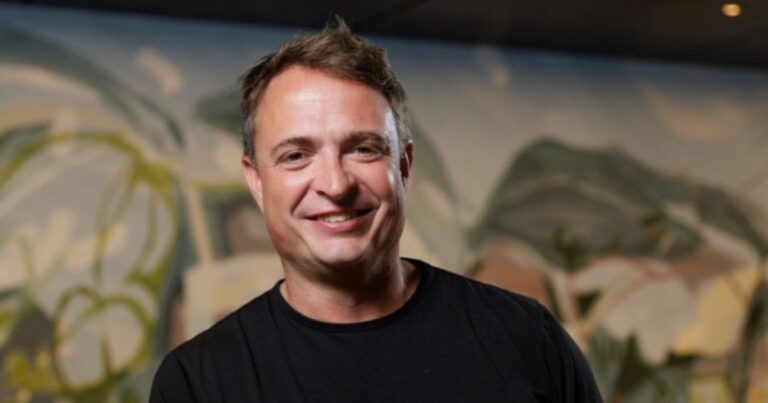 Pessoa vestindo uma camiseta preta, posicionada em ambiente interno com um mural artístico colorido ao fundo. A pintura apresenta formas fluídas e tons suaves, dando um clima moderno e criativo à cena. (Certta)