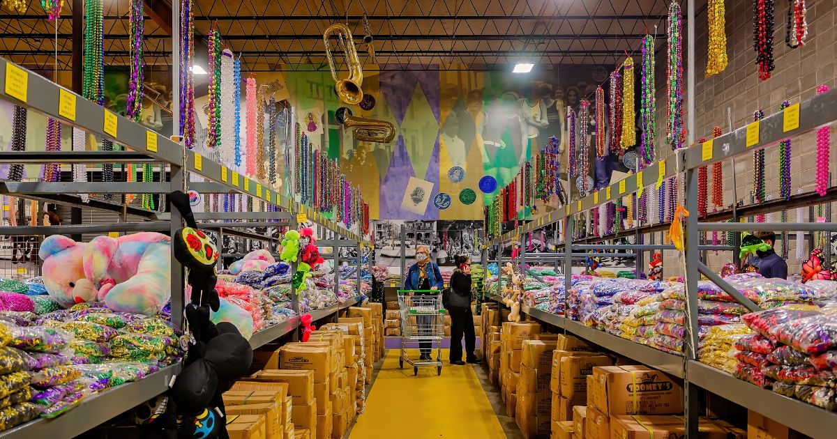 Um amplo depósito está repleto de itens coloridos típicos de festividades, especialmente colares de contas brilhantes dispostos em grandes quantidades em prateleiras metálicas altas. Pelúcias multicoloridas, máscaras e diversos acessórios festivos ocupam as laterais. No centro do corredor, duas pessoas com carrinhos de compras organizam ou selecionam produtos. O ambiente é iluminado e decorado com grandes murais nas paredes, exibindo instrumentos musicais dourados e figuras estilizadas, evocando o clima vibrante de celebrações como o Mardi Gras ou o Carnaval. Numerous caixas de papelão estão empilhadas no chão, indicando estoque volumoso.