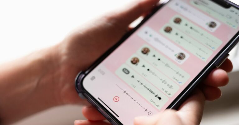 Uma pessoa segura um smartphone que exibe uma tela de aplicativo de mensagens. Diversas conversas estão abertas, mostrando múltiplos áudios enviados e recebidos, representados por ícones de reprodução e barras de ondas sonoras. O plano está focado na tela, enquanto as mãos que seguram o aparelho aparecem parcialmente desfocadas ao fundo. A interface do aplicativo tem fundo claro em tom rosado. (golpe)
