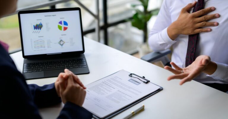 Duas pessoas estão sentadas em uma mesa durante uma conversa entre profissionais. Sobre a mesa, há um currículo preso a uma prancheta, uma caneta e um laptop exibindo gráficos coloridos de desempenho. Uma das pessoas está com as mãos entrelaçadas, enquanto a outra gesticula com uma mão no peito, sugerindo uma explicação ou resposta durante uma entrevista ou reunião de negócios.