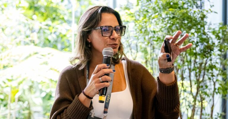 Uma pessoa está falando em um ambiente iluminado por luz natural, segurando um microfone em uma mão e um controle remoto de apresentação na outra. Ela veste um cardigã marrom sobre uma blusa clara e usa óculos. Ao fundo, há plantas verdes que compõem um cenário natural e suave. (mudança)