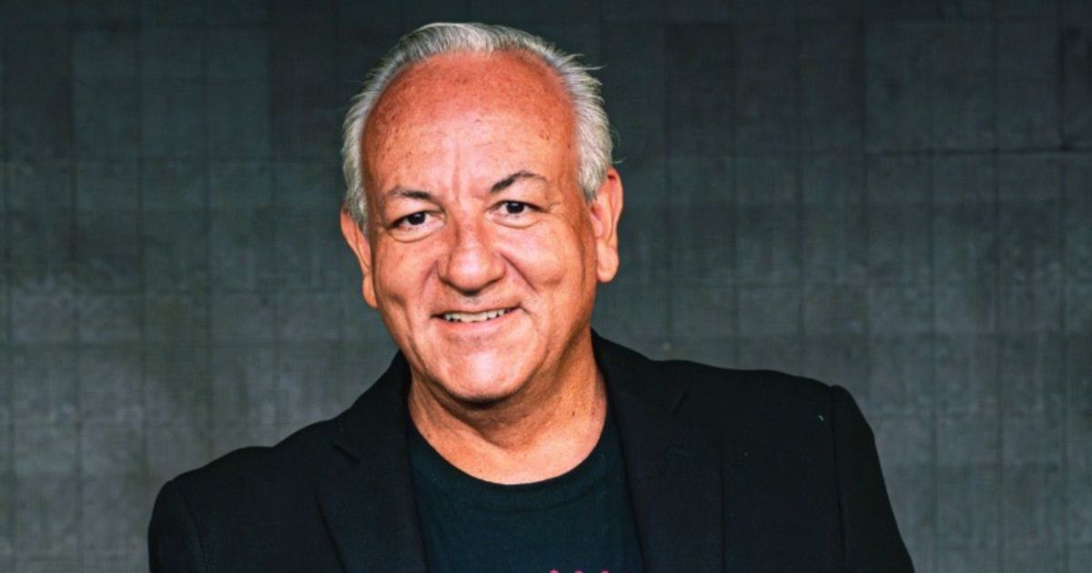 Uma pessoa de cabelos grisalhos veste uma camiseta preta sob um blazer escuro. Ela está posicionada diante de uma parede texturizada em tom cinza, em um ambiente com iluminação suave. A composição é centrada no tronco e nos ombros, transmitindo um estilo casual‑profissional. (F5)