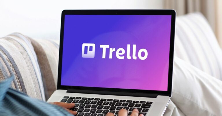 Uma pessoa utiliza um laptop apoiado no colo enquanto a tela exibe o logotipo do Trello sobre um fundo em degradê roxo‑azulado. O ambiente parece confortável, com almofadas claras ao redor, sugerindo um uso casual da ferramenta de produtividade em um espaço doméstico ou relaxado.