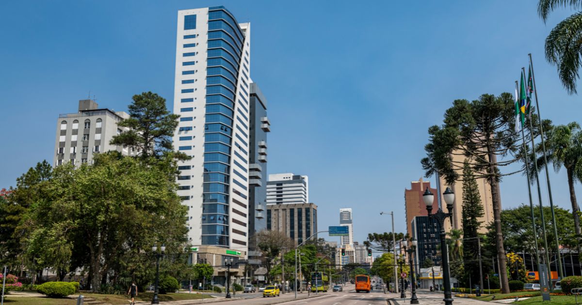 Vista urbana de uma avenida larga com prédios altos ao fundo em um dia ensolarado. À esquerda, há áreas verdes com árvores e gramados. No centro e à direita, aparecem edifícios modernos, postes de iluminação e bandeiras hasteadas. Um ônibus laranja circula pela via, que está com pouco movimento. (startups)