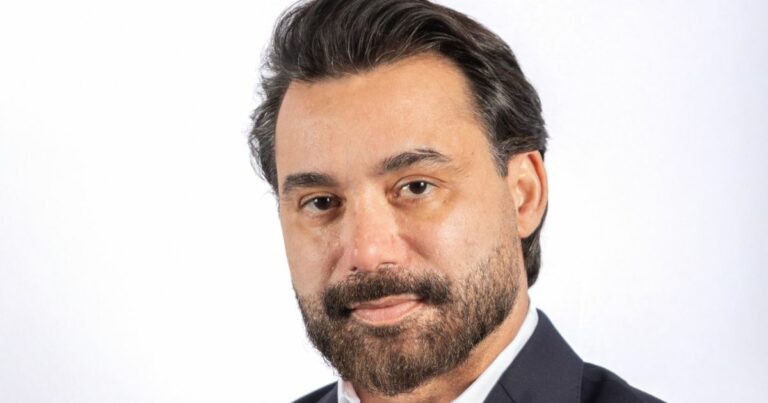 Uma pessoa vestindo um terno escuro com camisa branca aparece em um retrato profissional diante de um fundo totalmente branco. A imagem é enquadrada da altura do peito para cima, destacando o traje formal e o cabelo escuro cuidadosamente arrumado. (Approach Tech)