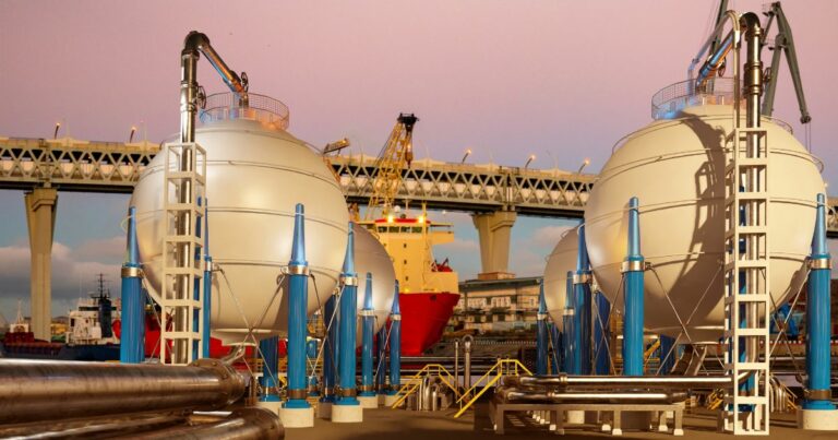 Estrutura industrial com grandes tanques esféricos brancos sustentados por colunas azuis, dispostos em uma área externa com tubulações metálicas em primeiro plano. Ao fundo, um navio vermelho e amarelo está atracado próximo a uma ponte elevada com múltiplos pilares, sob um céu em tons rosados de fim de tarde. (hidrogênio)