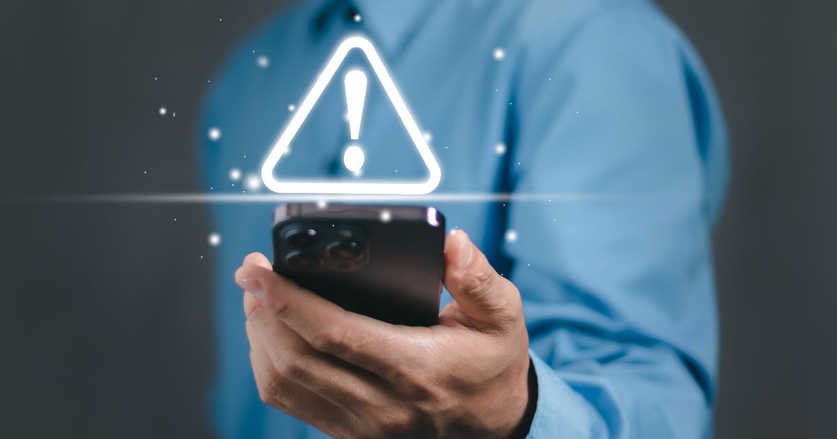 Uma pessoa usando camisa azul segura um smartphone na mão enquanto um ícone luminoso de alerta — um triângulo com ponto de exclamação — aparece projetado acima do aparelho, sugerindo uma notificação importante ou aviso digital. O fundo é neutro e desfocado, destacando o gesto e o símbolo de alerta. (vazamento)