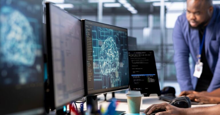 Várias telas de computador estão dispostas sobre uma mesa em um ambiente corporativo moderno. O monitor principal exibe a imagem de um cérebro estilizado composto por circuitos, sugerindo visualizações de inteligência artificial ou redes neurais. Ao lado, outro monitor mostra linhas de código. Uma pessoa digita no teclado enquanto outra, usando crachá, se inclina sobre a mesa analisando as telas. Sobre a mesa também há um fone de ouvido, uma xícara descartável e outros equipamentos de trabalho. O fundo inclui painéis de vidro que deixam o ambiente iluminado. (capital)