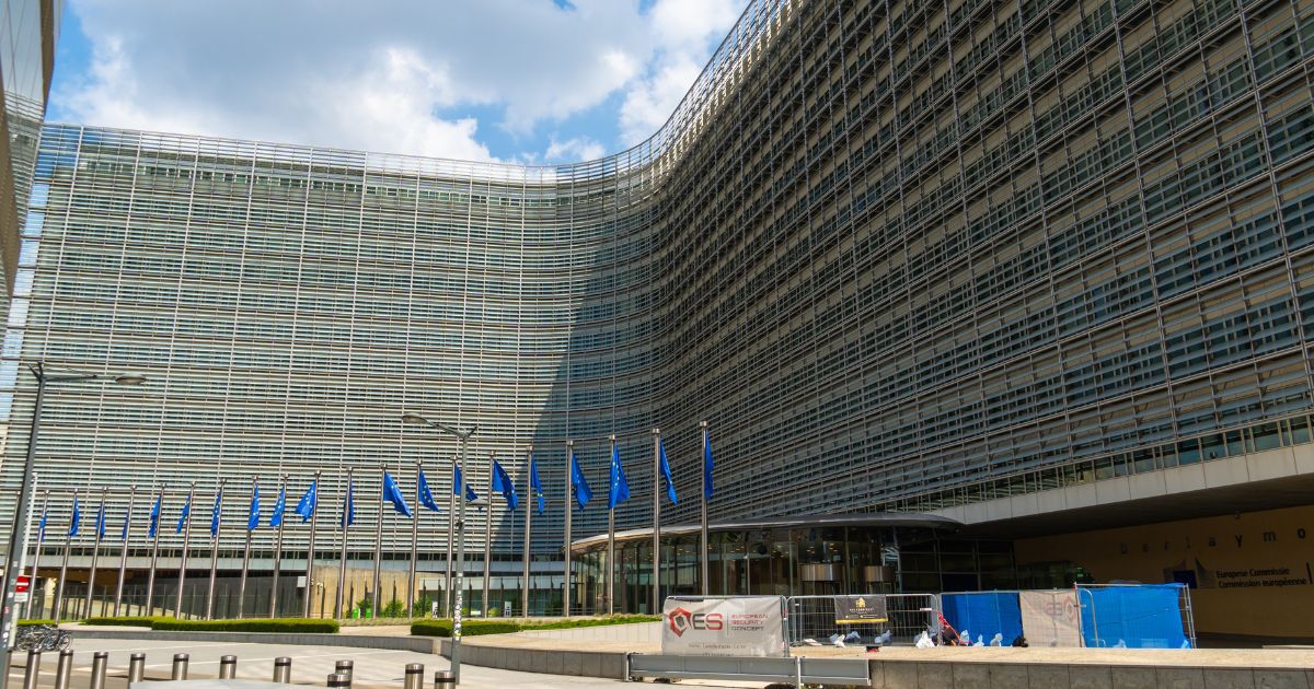 Uma grande edificação moderna com fachada curva e composta por múltiplas fileiras de estruturas metálicas e janelas domina a cena. Em frente ao prédio, há uma sequência de mastros exibindo bandeiras azuis com estrelas amarelas. O céu está parcialmente nublado, e algumas barreiras e equipamentos de obra aparecem próximos à entrada, sugerindo manutenção ou instalação temporária no local. (nuvem soberana)