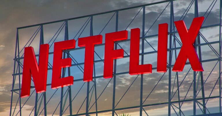 Um grande letreiro tridimensional com a palavra “NETFLIX” em letras vermelhas está instalado sobre uma estrutura metálica elevada. O céu ao fundo exibe tons de azul, cinza e laranja, sugerindo um pôr do sol ou amanhecer, enquanto a iluminação realça as letras vermelhas brilhantes do logotipo.