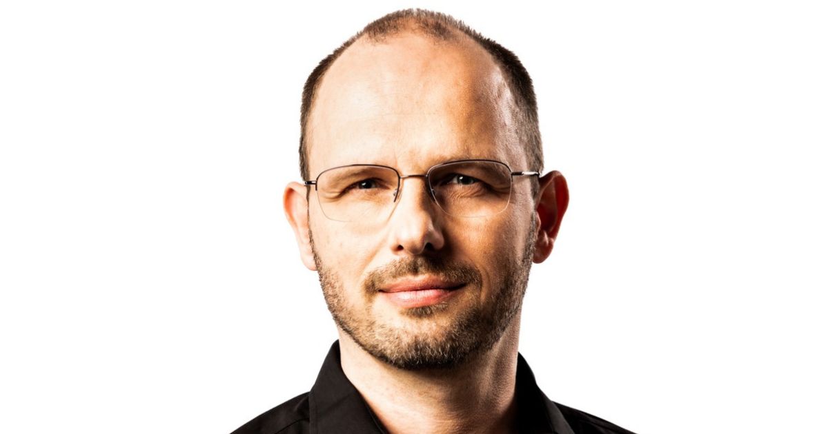 Uma pessoa vestindo uma camisa preta aparece em um retrato com fundo totalmente branco. A imagem é enquadrada da altura dos ombros para cima, destacando a gola da camisa e a postura frontal. (Github)