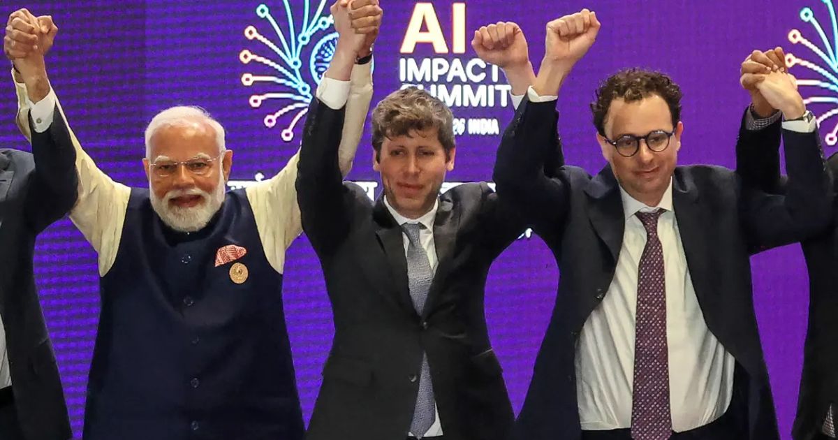 Um grupo de pessoas em trajes formais está de pé em um palco, com os braços erguidos e as mãos dadas em sinal de celebração ou união. Elas usam ternos escuros, camisas sociais e gravatas. Ao fundo, um grande painel roxo exibe o texto “AI Impact Summit”, acompanhado de elementos gráficos decorativos em formato de explosões luminosas estilizadas, criando um clima de evento tecnológico ou conferência internacional. (Anthropic)