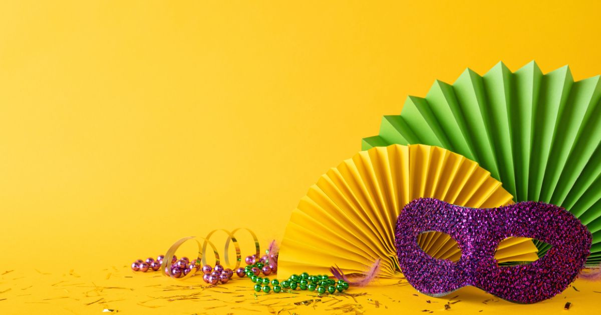 Itens de festa coloridos estão dispostos sobre um fundo amarelo vibrante. À direita, há uma máscara roxa com glitter, e atrás dela dois leques de papel em amarelo e verde. No canto esquerdo inferior, colares de contas metálicas em tons de roxo, dourado e verde aparecem espalhados, junto com serpentinas e pequenos confetes. A composição transmite clima de celebração, típico de festividades como Carnaval. (bloco)