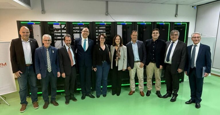 Um grupo de dez pessoas posa lado a lado em um ambiente interno com piso verde-claro. Elas vestem trajes formais e semiformais, como ternos, camisas sociais, blazers e gravatas. Ao fundo, há uma fileira de racks de servidores iluminados com luzes verdes nas bordas, indicando um ambiente tecnológico ou de infraestrutura de TI. À esquerda, parte de um banner institucional é visível. A composição sugere uma visita oficial, apresentação ou cerimônia relacionada a tecnologia ou centros de dados. (USP)