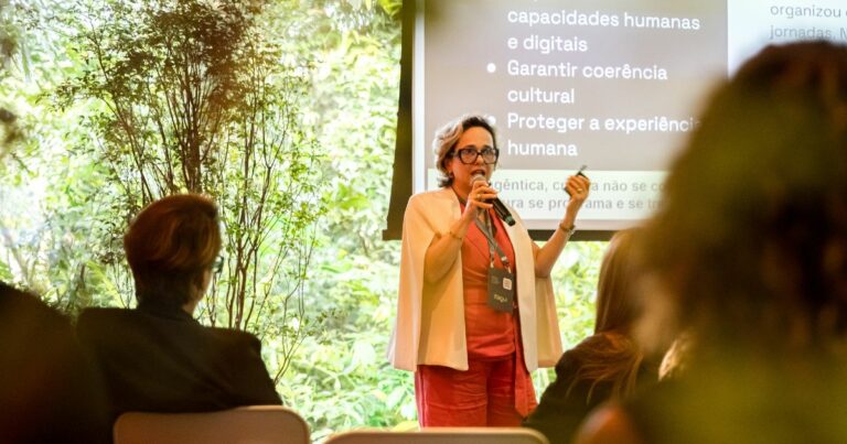 Uma pessoa em pé, usando roupas claras e segurando um microfone, faz uma apresentação diante de um pequeno público. Ela está posicionada entre um ambiente externo com vegetação ao fundo e um telão que exibe texto relacionado a capacidades humanas, coerência cultural e experiência humana. O público aparece sentado em primeiro plano, observando a apresentação. (governança)