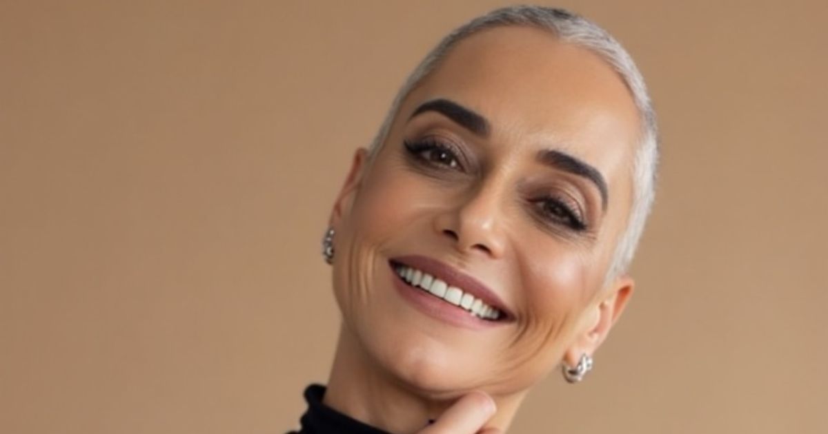 Uma pessoa com cabelo curto e grisalho aparece em um retrato posado diante de um fundo bege uniforme. Ela veste uma blusa de gola alta preta e apoia a mão no queixo, em um gesto contemplativo. Um brinco metálico é visível em uma das orelhas, e a composição tem estilo minimalista e elegante. (filosofias)